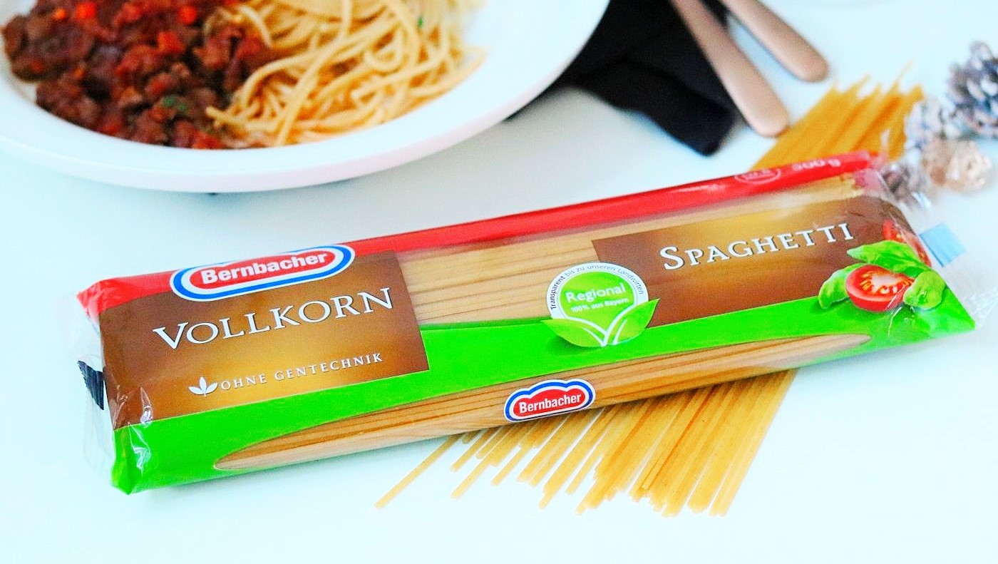 Nudelpackung, Bernbacher Vollkornspaghetti auf weißem Grund, im Hintergrund Teller mit Spaghetti und Bolognese