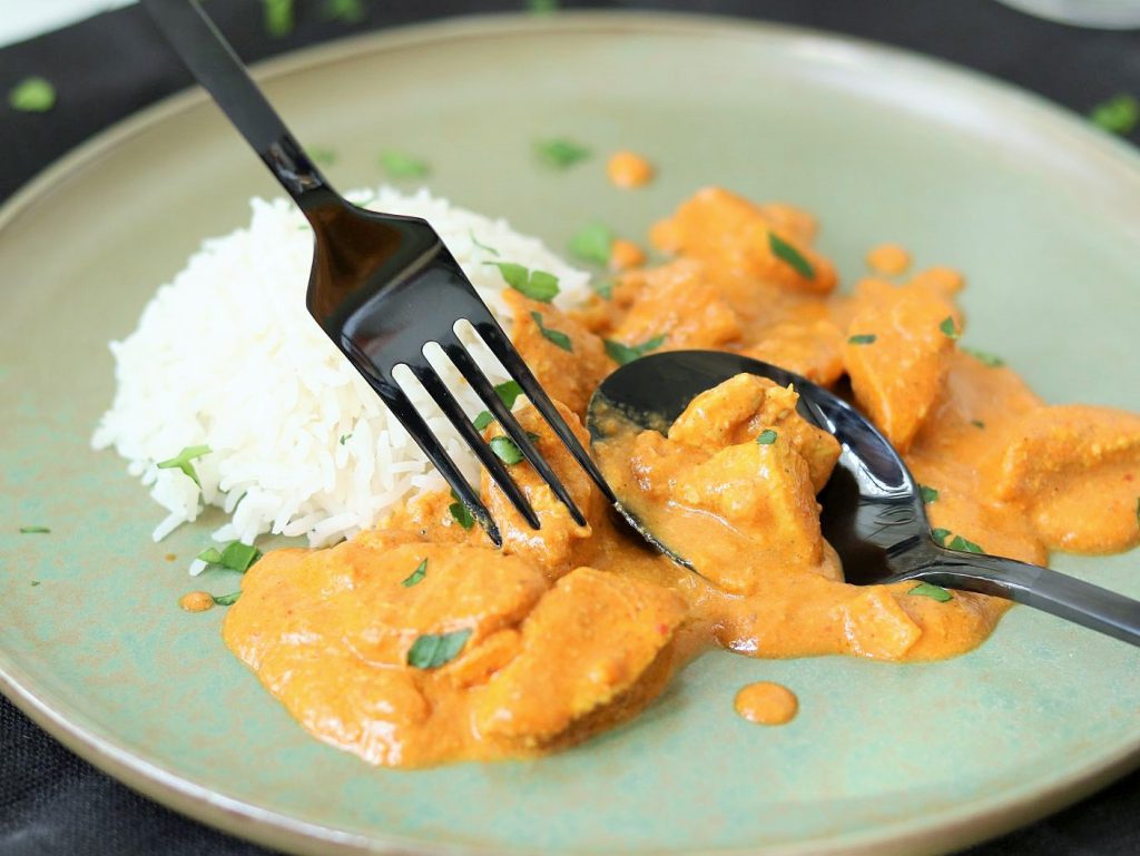 Hellgrüner Teller, darauf orange farbenes Butter Chicken und weißer Reis, eine Gabel die in ein Fleischstück sticht