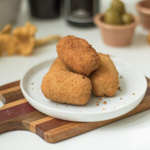 Tapas: vier Croquetas auf einem weißen Teller, im Hintergrund Oliven und Pfifferlinge