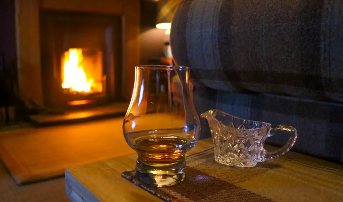 Schottland Roadtrip, Glas Whisky auf einem Holztisch vor einem Kamin mit Feuer