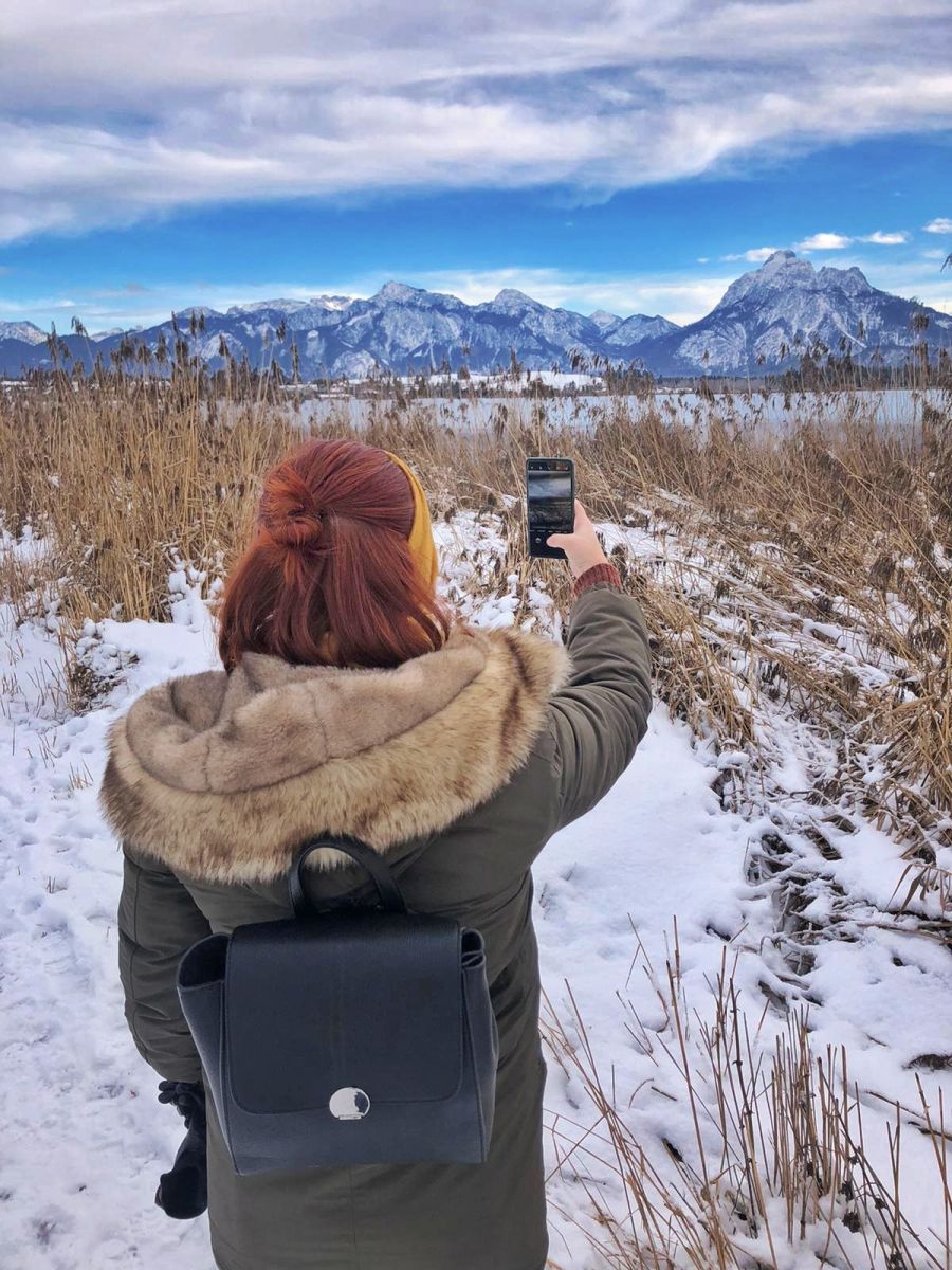 Winterspaziergang am Hopfensee in Bayern, eine Frau fotografiert mit dem Handy das Alpenpanorama am Seeufer