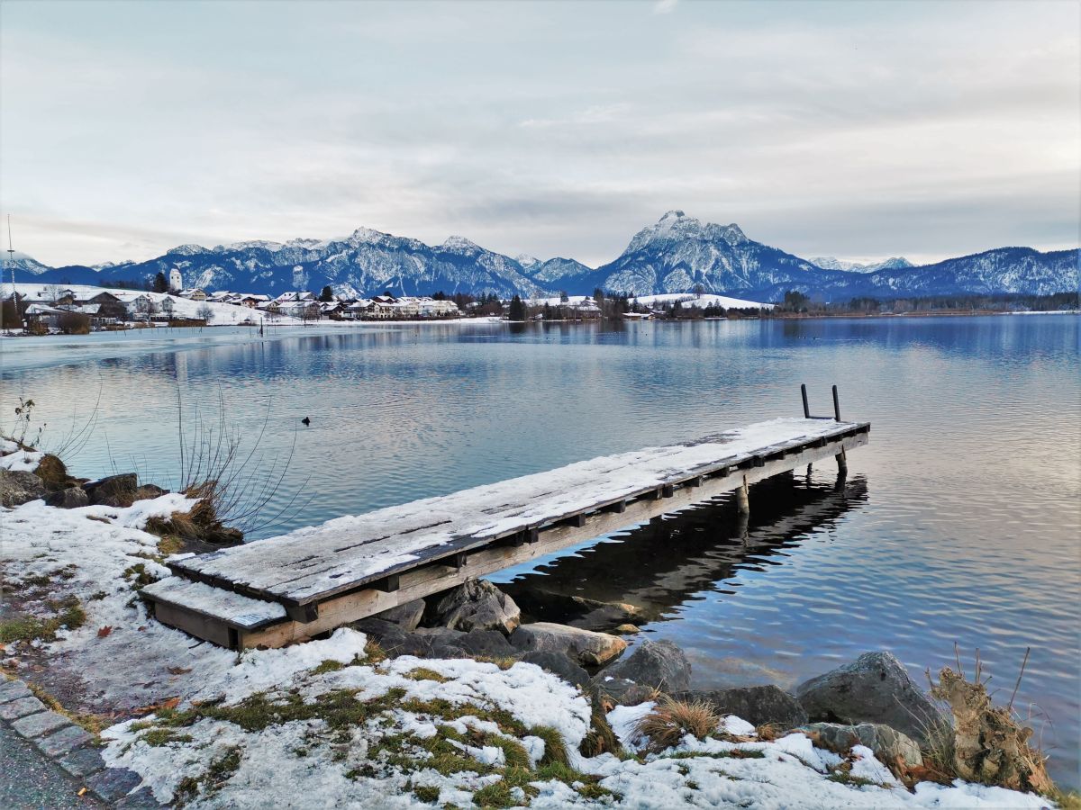 Winterspaziergang am Hopfensee in Bayern, Blick auf einen Schnee bedeckten Steg am Seeufer mit Blick auf die Allgäuer Berge