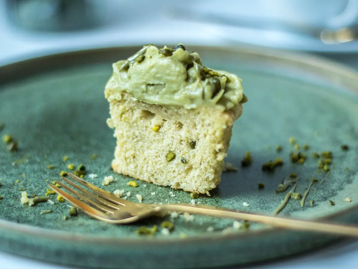 Ein angeschnittener Pistazien Cupcake auf einem grünen Teller, auf dem Teller liegt noch eine goldene Kuchengabel