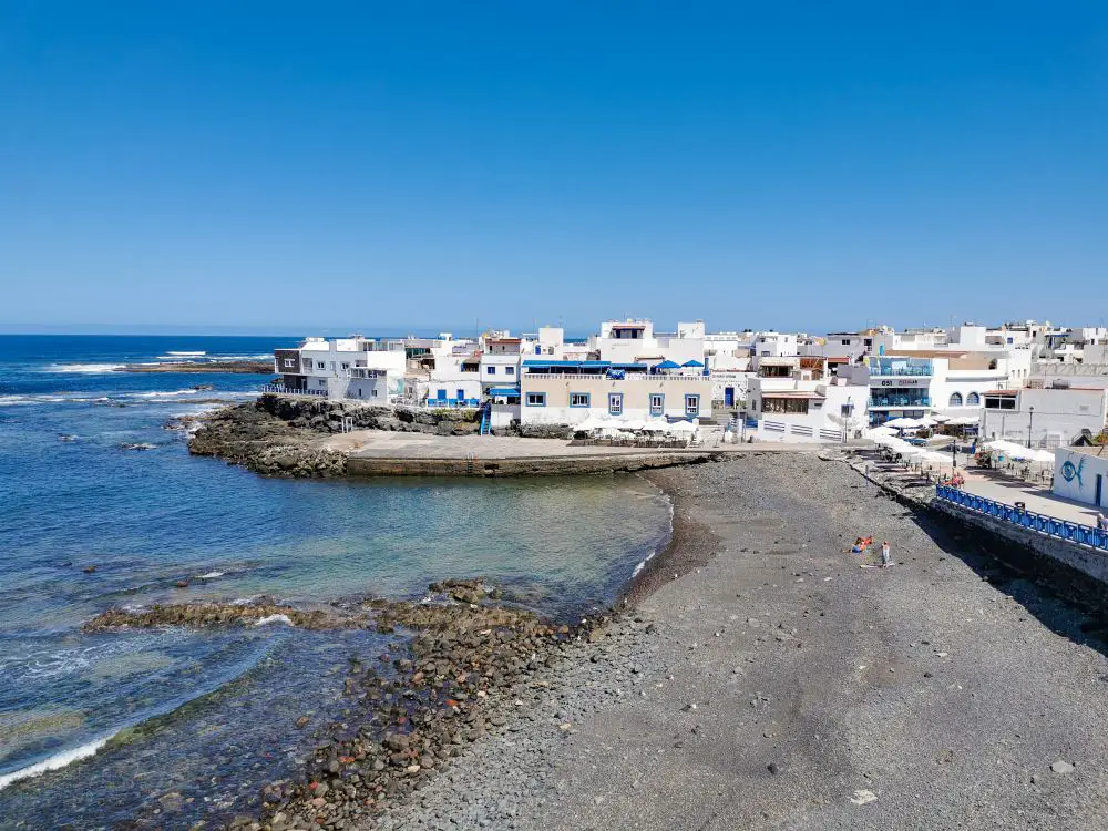 Fischerdorf El Cotillo, eine Bucht mit kleinem Strand, weiße Häuser mit blauen Sonnenschirmen.