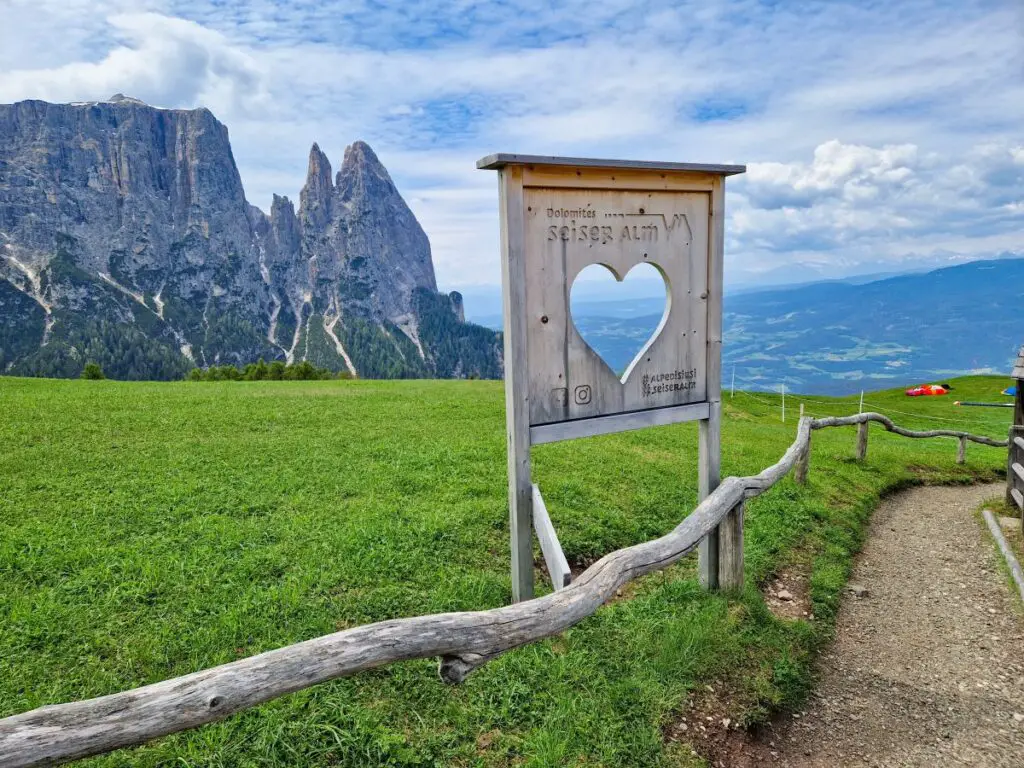 Holztafel mit ausgeschnittenem Herz als Fenster, dahinter die Berge der Dolomiten